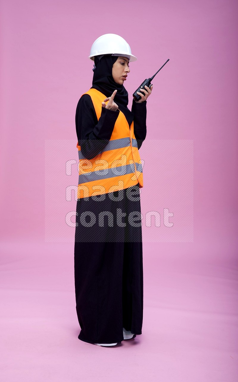 Saudi woman wearing Abaya with engineer vest and helmet standing holding walkie-talkie on pink background