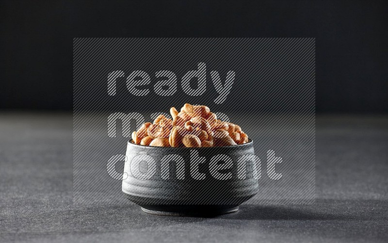 A black pottery bowl full of cashews on a black background in different angles