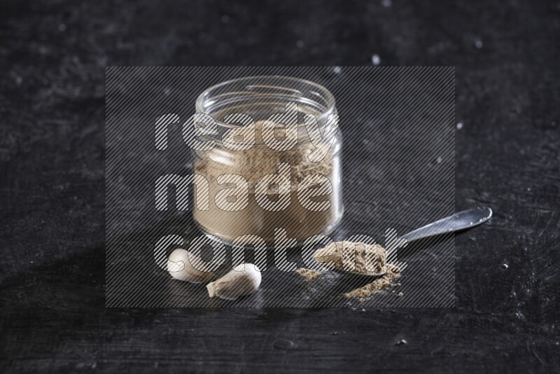 A glass jar full of garlic powder with a metal spoon full of the powder on a textured black flooring