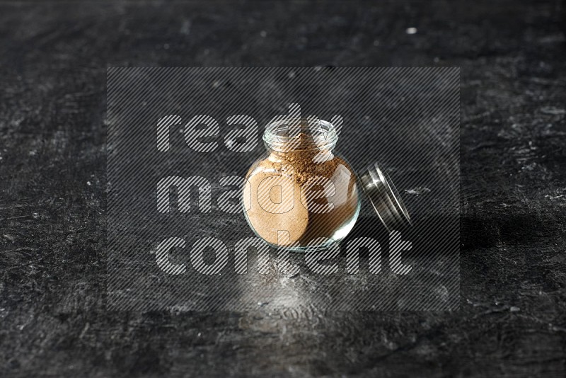 A glass spice jar full of allspice powder on a textured black flooring