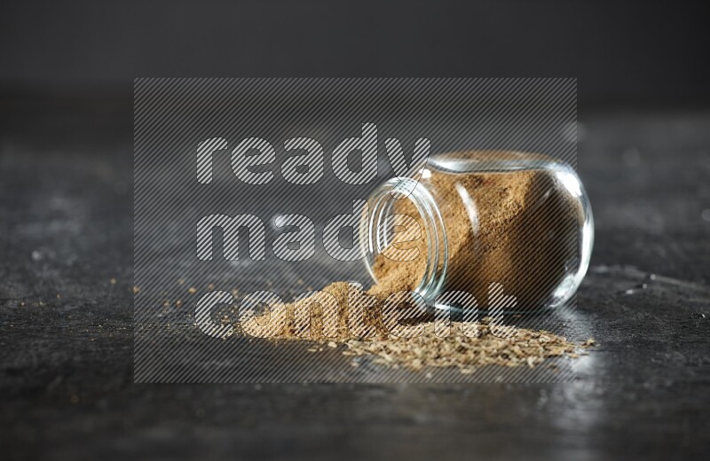 A flipped glass spice jar full of cumin powder with spilled powder and cumin seeds on a textured black flooring