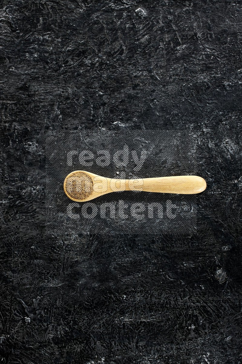 A wooden spoons full of cumin powder on textured black flooring