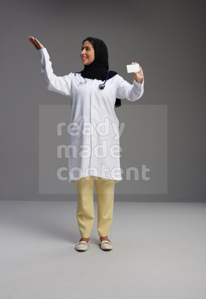 Saudi woman wearing lab coat with stethoscope standing holding ATM card on Gray background