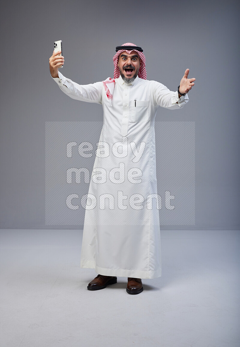 Saudi man Wearing Thob and red Shomag standing taking selfie on Gray background