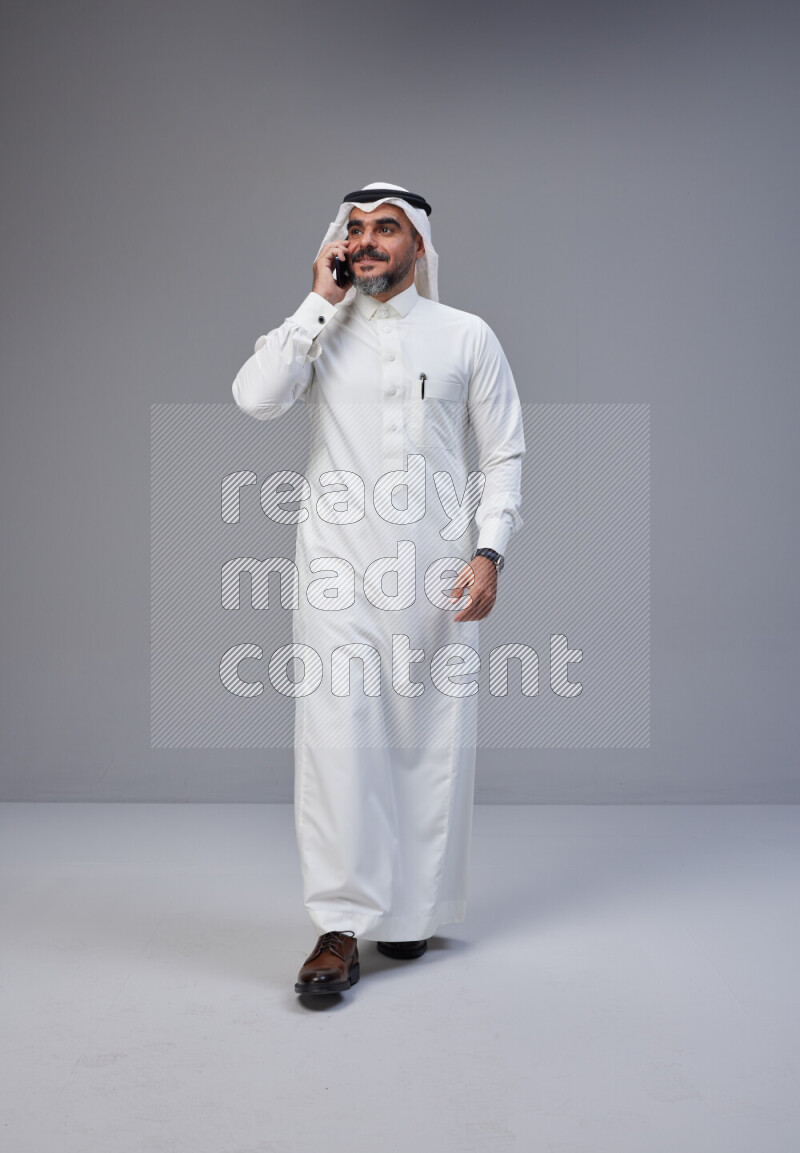 Saudi man Wearing Thob and white Shomag standing talking on phone on Gray background