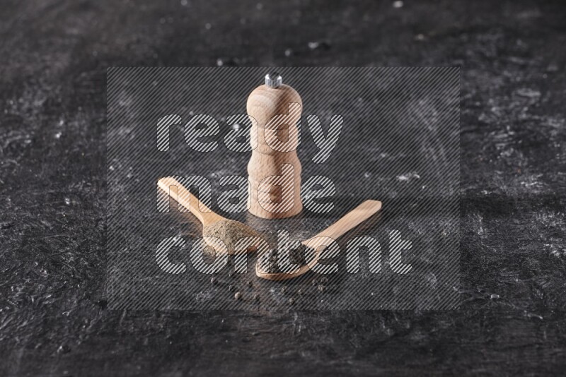 Black pepper powder and black pepper beads in wooden spoons and wooden grinder on a textured black flooring