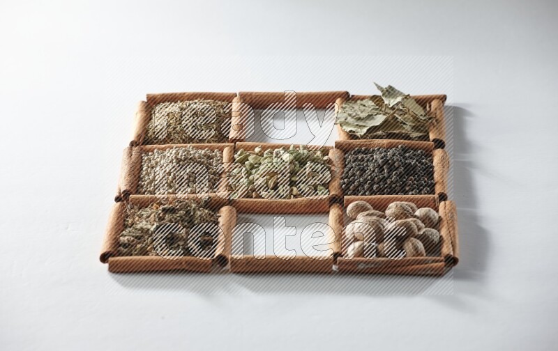 9 squares of cinnamon sticks full of cardamom in the middle surrounded by nutmeg, salt, dried basil, white pepper, cumin, allspice, bay laurel leaves and black pepper on white flooring