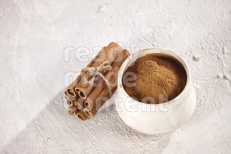 Cinnamon sticks stacked and bounded beside a beige bowl full of cinnamon powder on white background