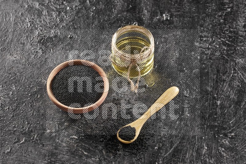 وعاء خشبي وملعقة ممتلئان ببذور حبة البركة وجرة زجاجية من زيت حبة البركة على أرضية سوداء