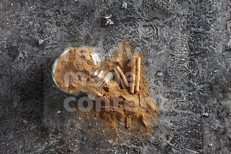 Flipped glass herbs jar full of cinnamon powder with cracked cinnamon sticks on a textured black background