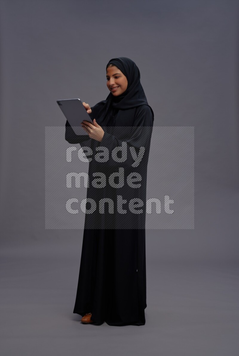 Saudi woman wearing Abaya standing working on tablet on gray background