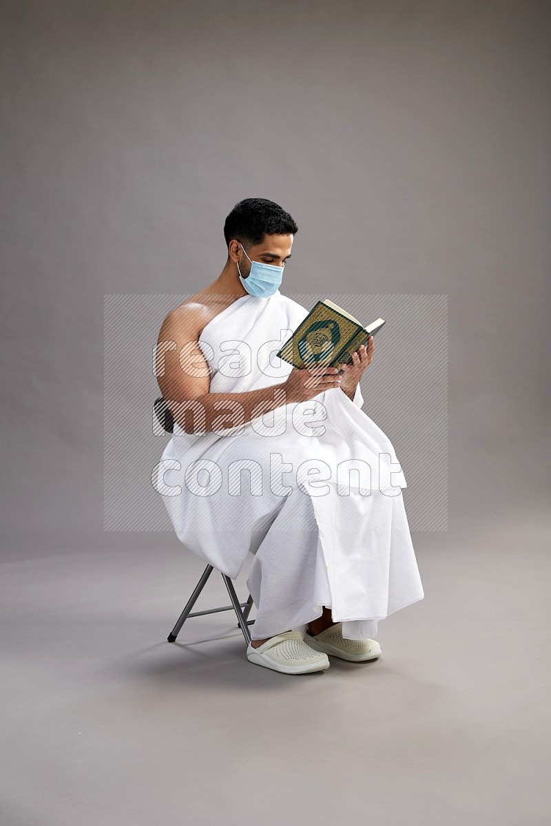 A man wearing Ehram with face mask sitting on chair reading quran on gray background