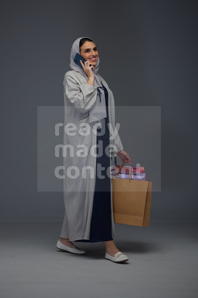 A Saudi woman wearing a light gray Abaya and head scarf standing and holding shopping bags making different poses eye level on a grey background