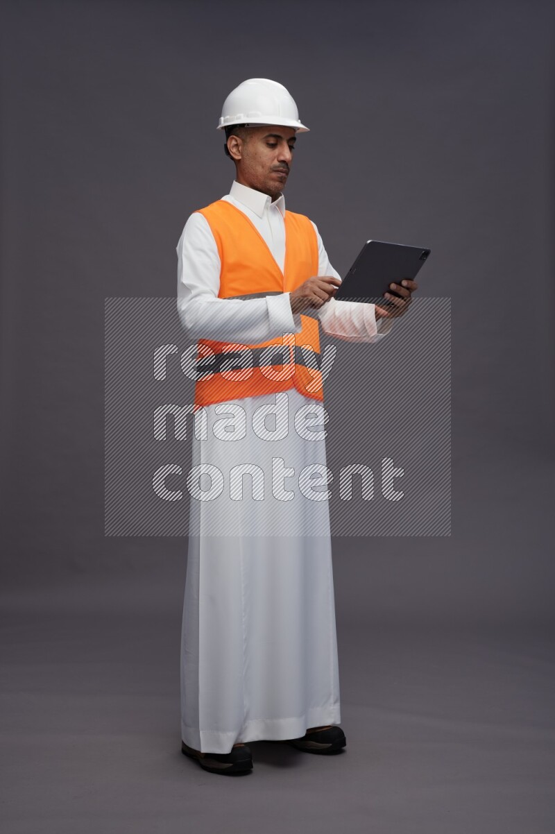 Saudi man wearing thob with engineer vest standing working on tablet on gray background