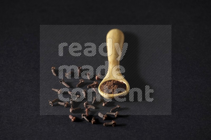 A wooden spoon full of cloves powder with spreaded whole cloves on a black flooring