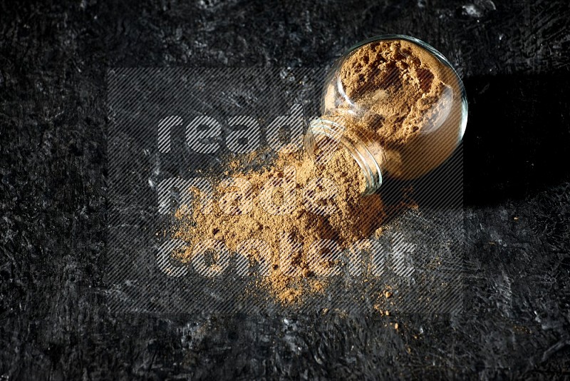 A flipped glass spice jar full of allspice powder and powder spilled out of it on a textured black flooring