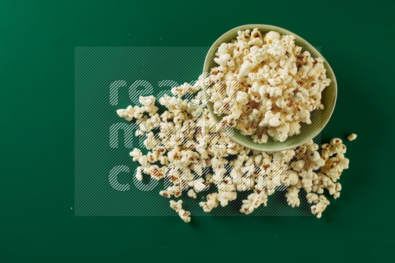 A green ceramic bowl full of popcorn with popcorn beside it on a green background in a top view shot