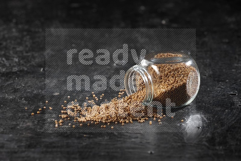A glass spice jar full of mustard seeds and jar is flipped with fallen seeds on a textured black flooring in different angles