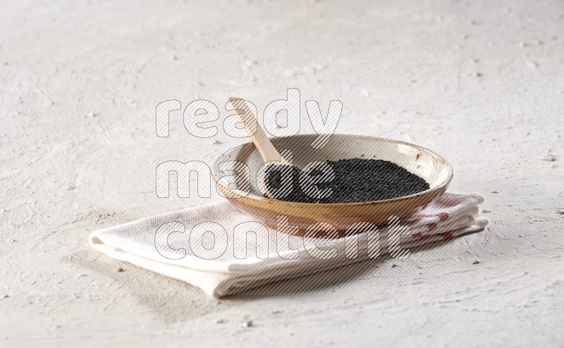 A multicolored pottery plate full of black seeds with a wooden spoon full of the seeds on a napkin on a textured white flooring