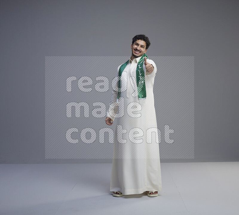 A Saudi man standing wearing thob and Saudi flag scarf on gray background