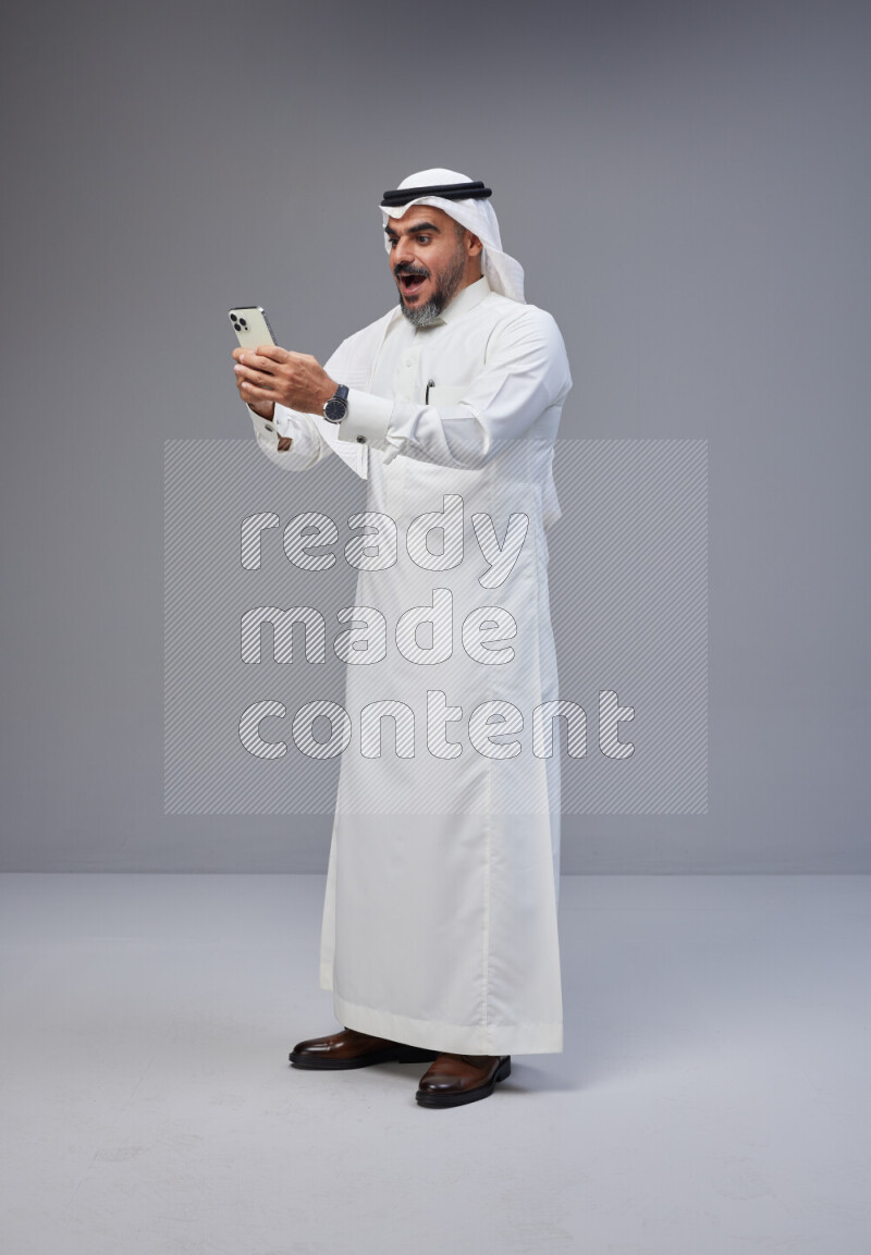 Saudi man Wearing Thob and white Shomag standing texting on phone on Gray background