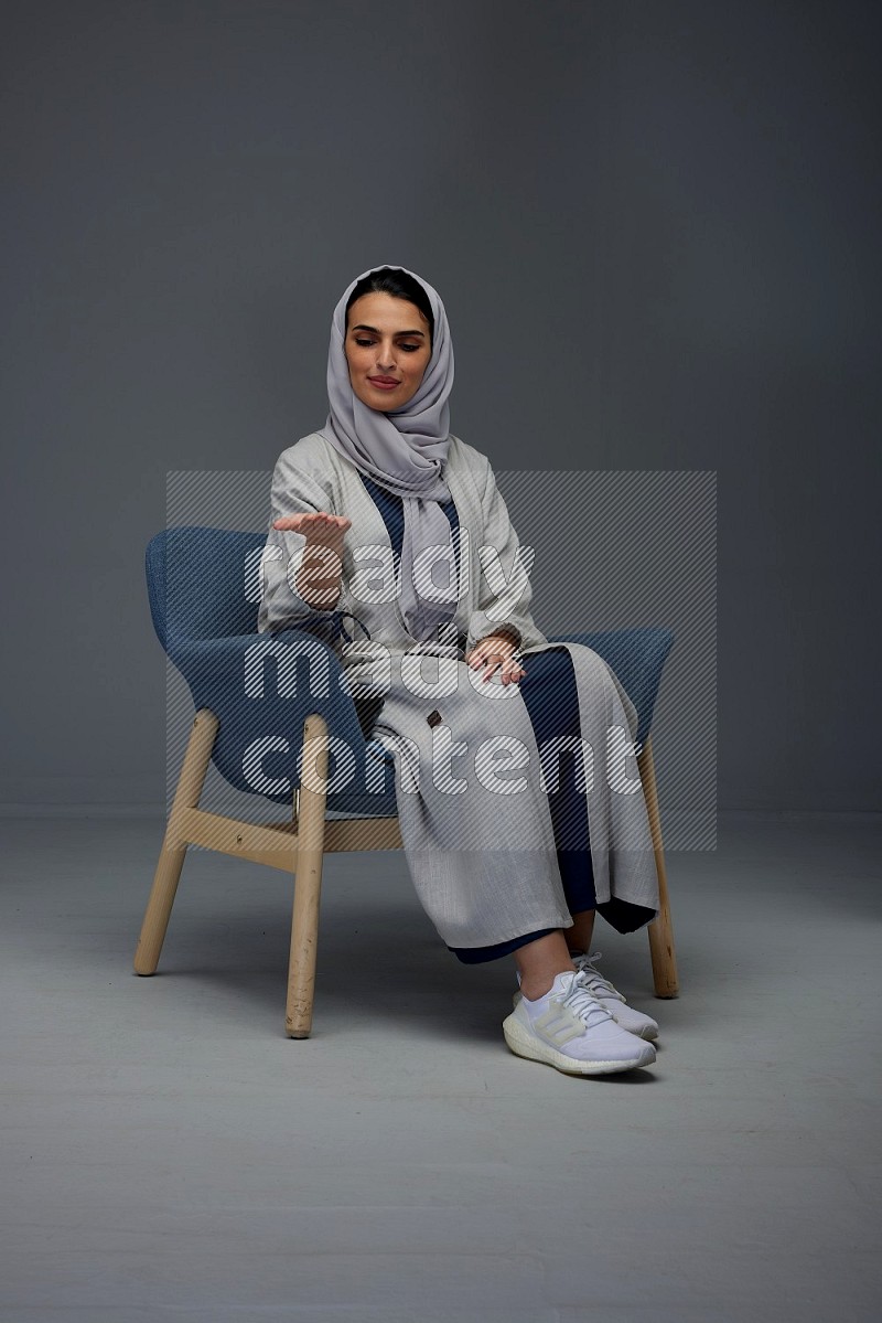 A Saudi woman wearing a light gray Abaya and head scarf sitting on a dark grey chair while making multi hand gestures eye level on a grey background