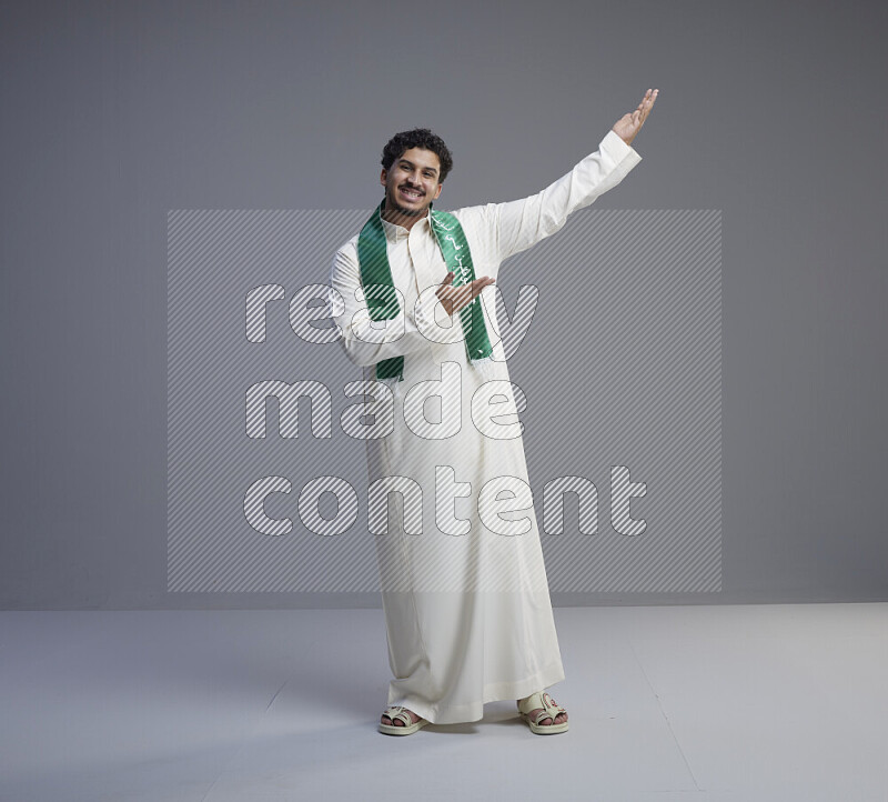 A Saudi man standing wearing thob and Saudi flag scarf on gray background