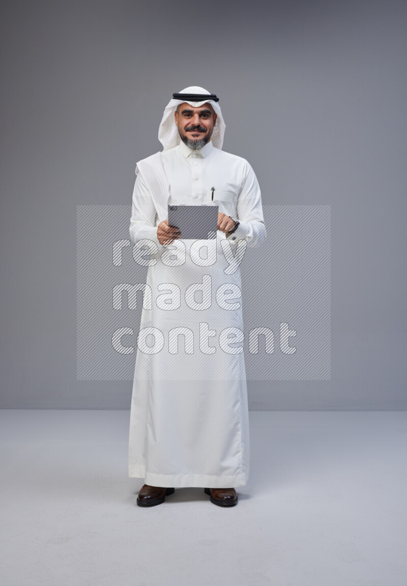 Saudi man Wearing Thob and white Shomag standing working on tablet on Gray background