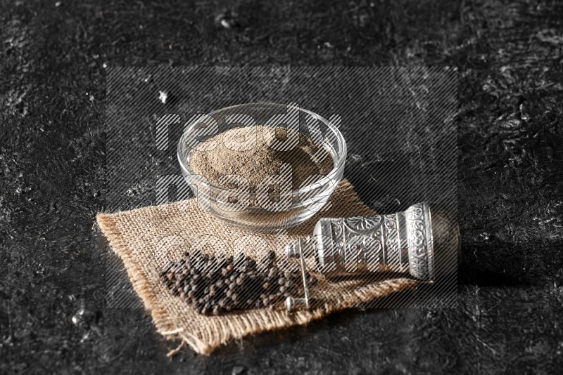 A glass bowl full of black pepper powder and black pepper beads on burlap fabric with a turkish metal pepper grinder on textured black flooring