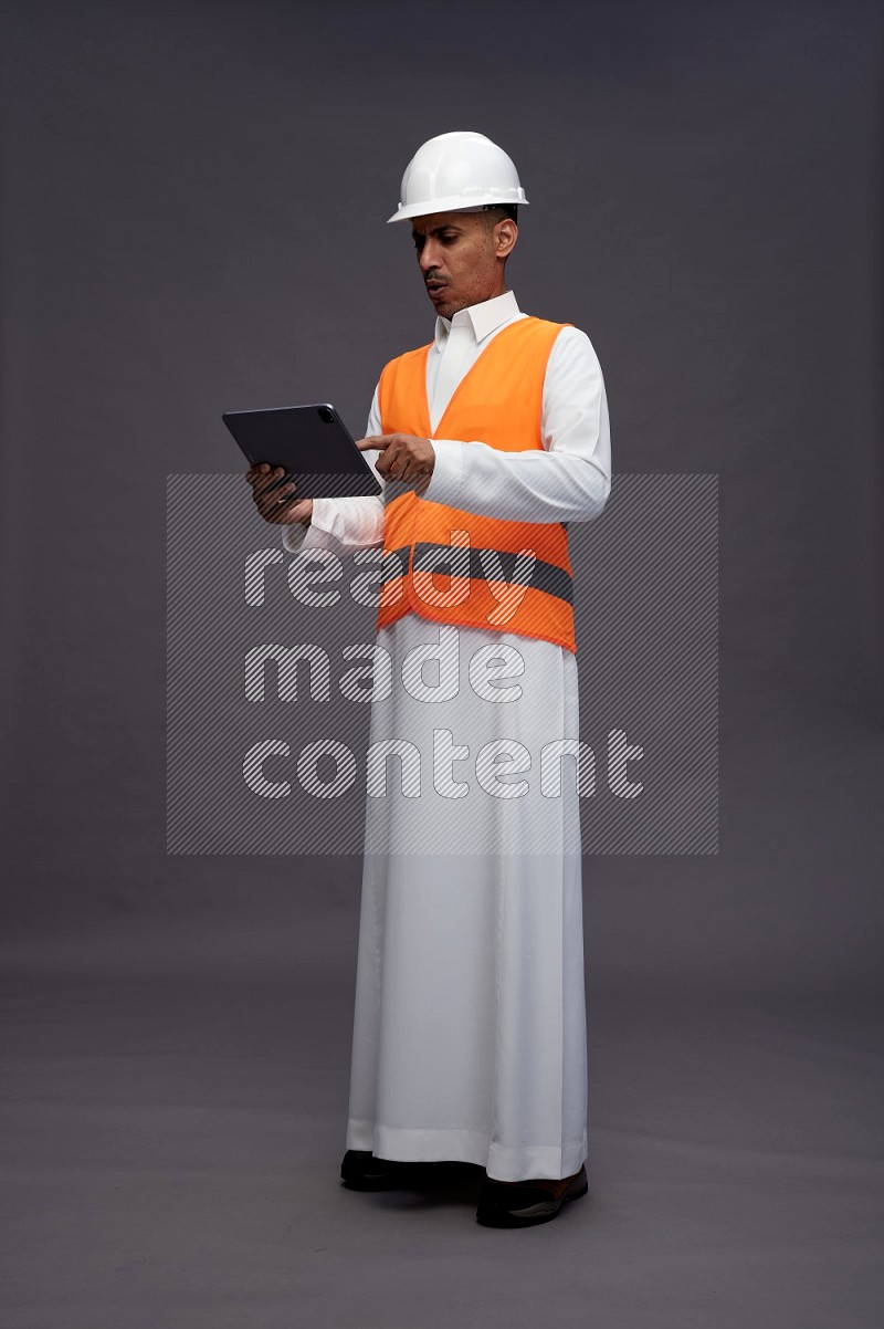 Saudi man wearing thob with engineer vest standing working on tablet on gray background