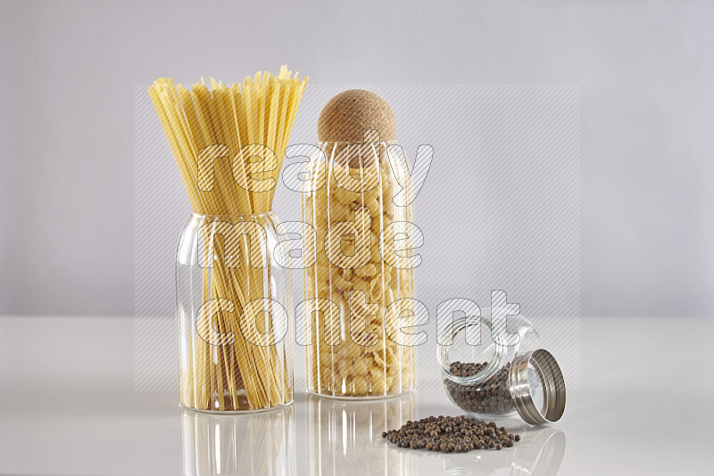 Raw pasta in glass jars with black peppers on light grey background