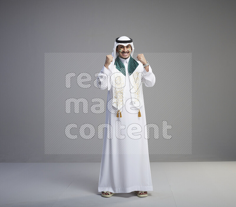 A Saudi man standing wearing thob and white shomag with face painting and Saudi flag scarf on gray background