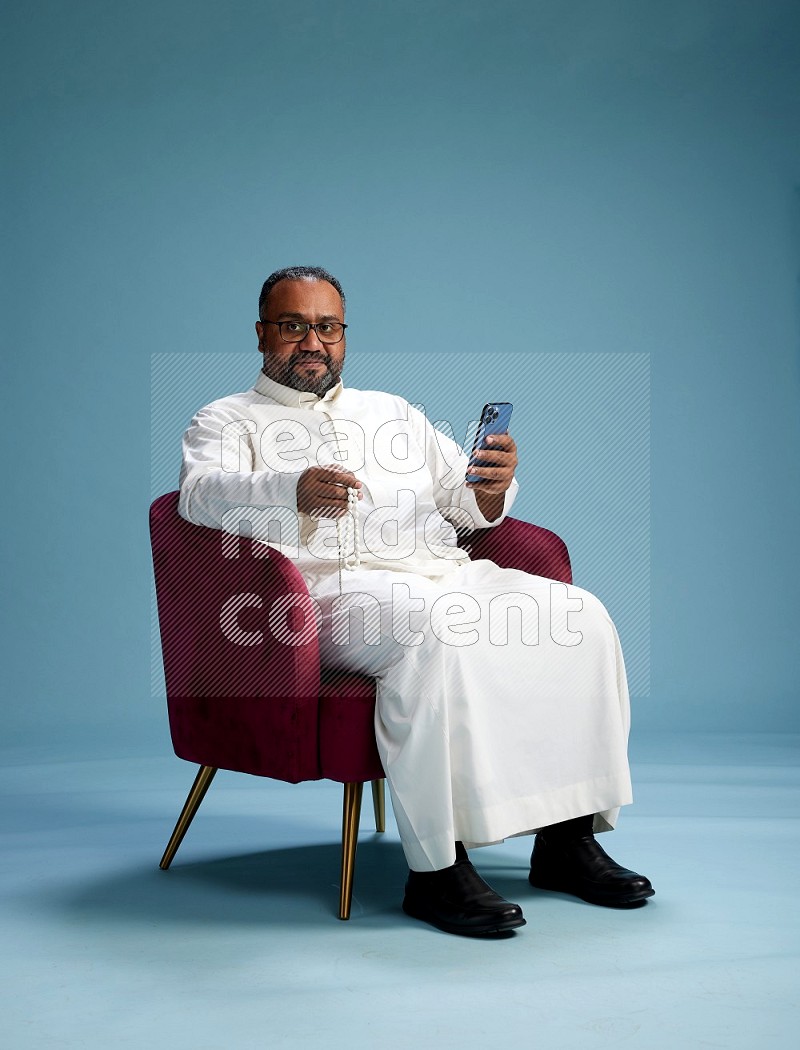 Saudi Man without shimag sitting on chair texting on phone on blue background