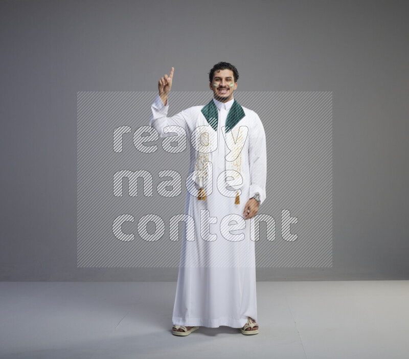 A Saudi man standing wearing thob and Saudi flag scarf with face painting on gray background