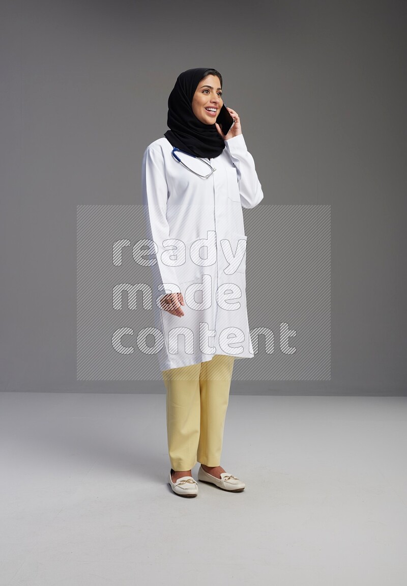 Saudi woman wearing lab coat with stethoscope standing talking on phone on Gray background