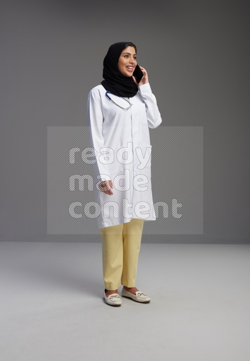 Saudi woman wearing lab coat with stethoscope standing talking on phone on Gray background
