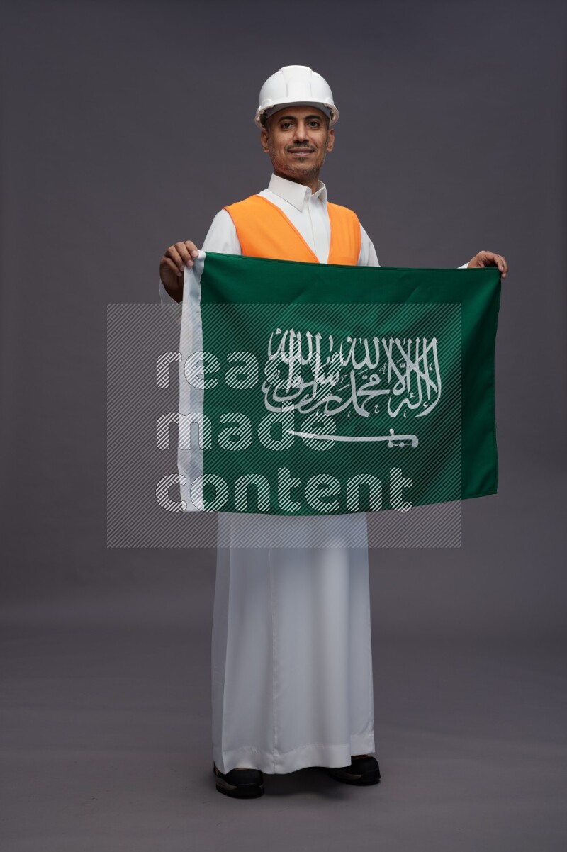 Saudi man wearing thob with engineer vest standing holding Saudi flag on gray background