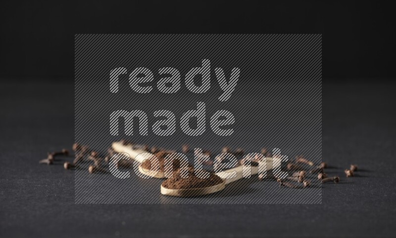 2 wooden spoons full of cloves powder with spreaded whole cloves on a black flooring