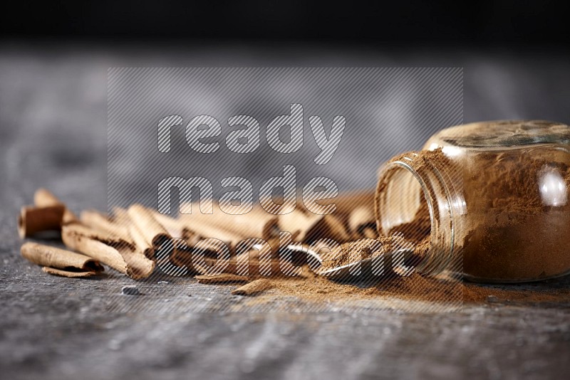 Herbal glass jar full cinnamon powder flipped and a metal spoon full of powder surrounded by cinnamon sticks on textured black background in different angles