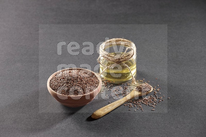 A wooden bowl and wooden spoon full of flaxseeds with a glass jar of flaxseeds oil on a black flooring