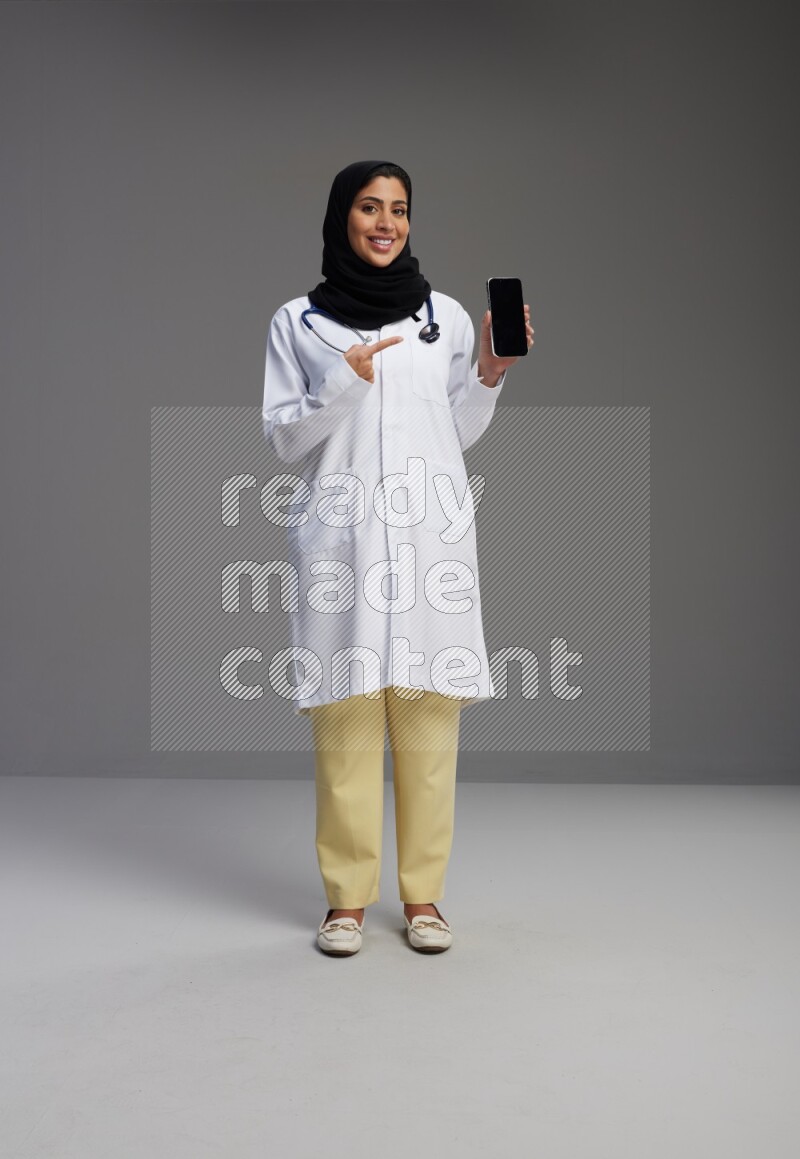 Saudi woman wearing lab coat with stethoscope standing showing phone to camera with sign in the back on Gray background