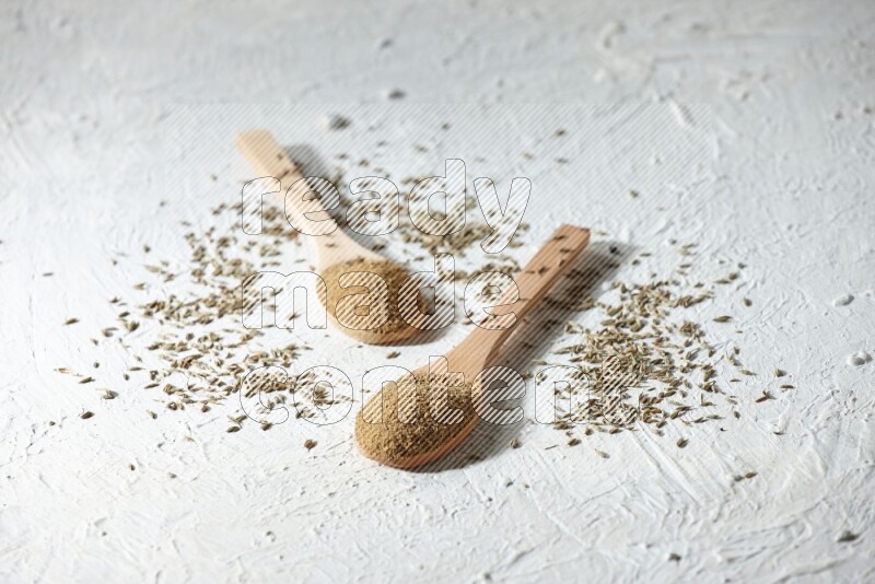 2 Wooden spoons full of cumin powder and cumin seeds on textured white flooring