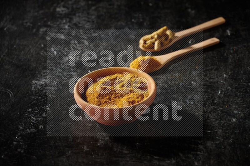 A wooden bowl full of turmeric powder and 2 wooden spoons full of dried turmeric finger and turmeric powder on textured black flooring