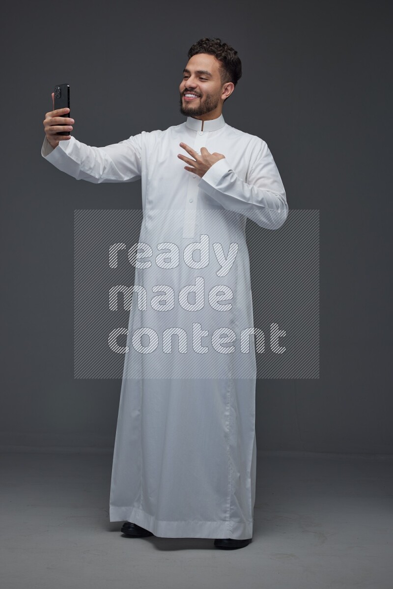 A Saudi man wearing Thobe and taking selfie with his phone making different poses eye level on a gray background