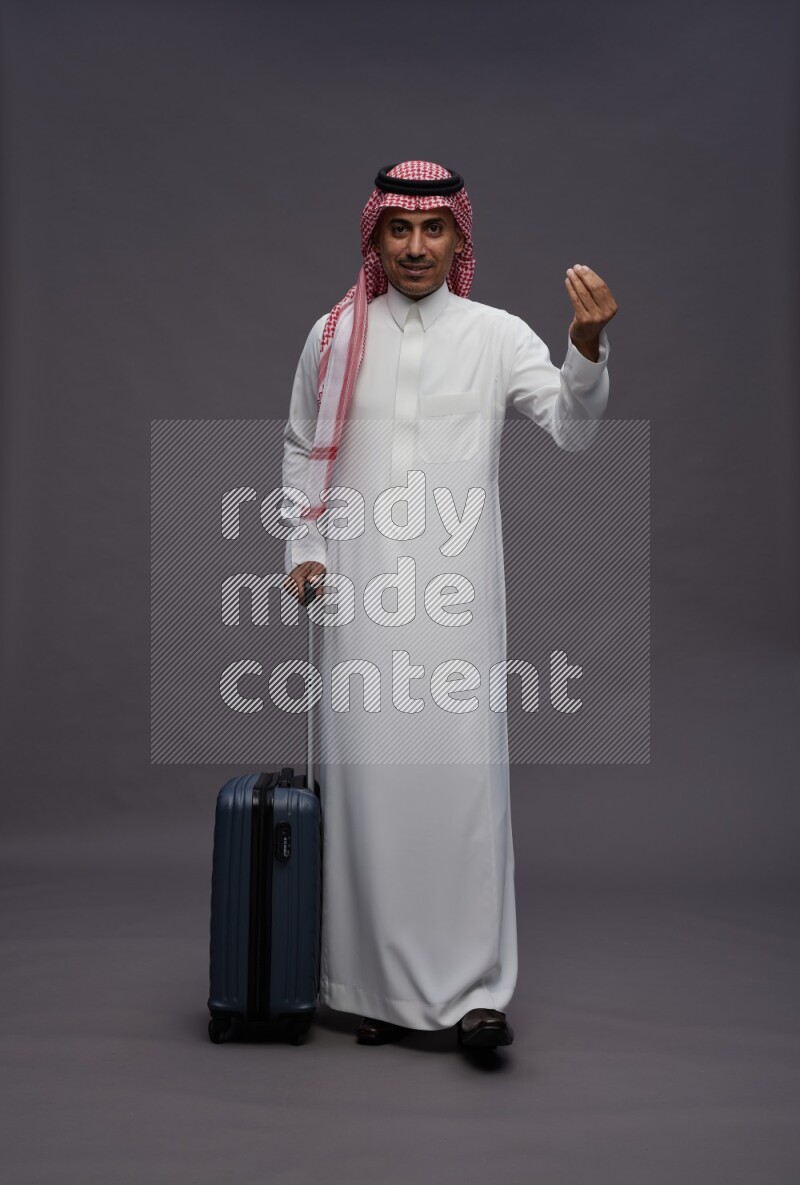 Saudi man wearing thob and shomag standing holding bag on gray background