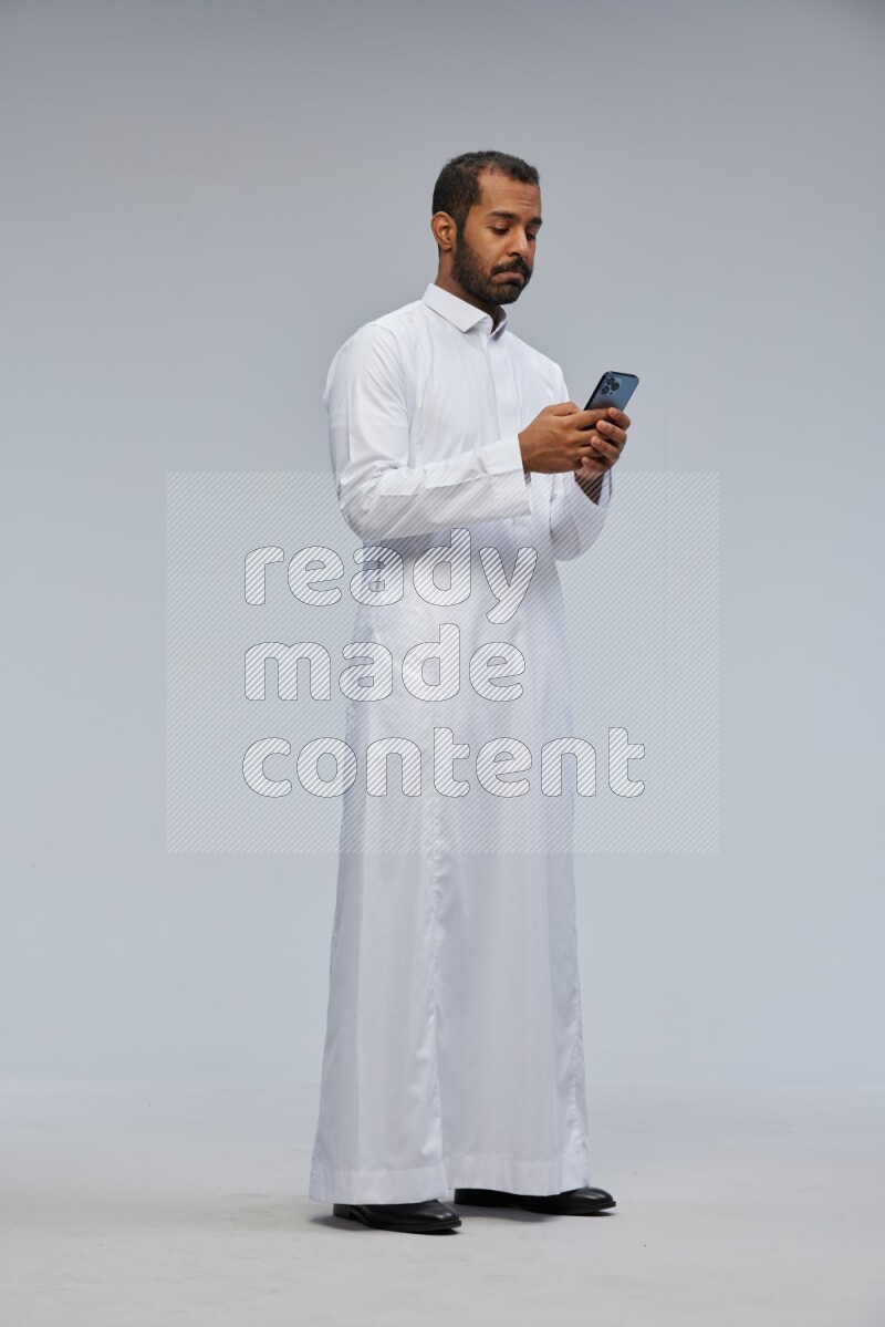 Saudi man wearing Thob standing texting on phone on Gray background