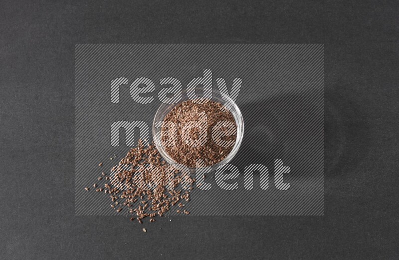 A glass bowl full of flaxseeds surrounded by the seeds on a black flooring