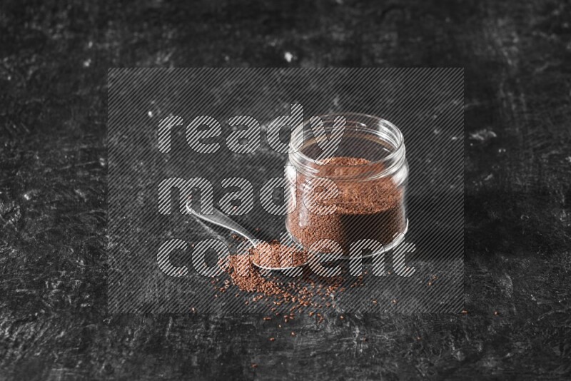 A glass jar full of garden cress seeds with a metal spoon full of the seeds on a textured black flooring