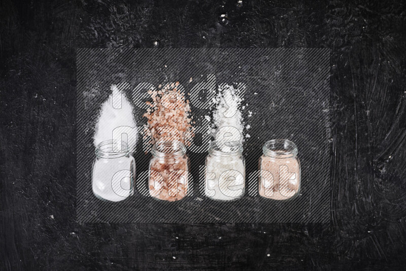 4 glass jars filled with table salt, coarse sea salt, fine himalayan salt and coarse himalayan salt on black background