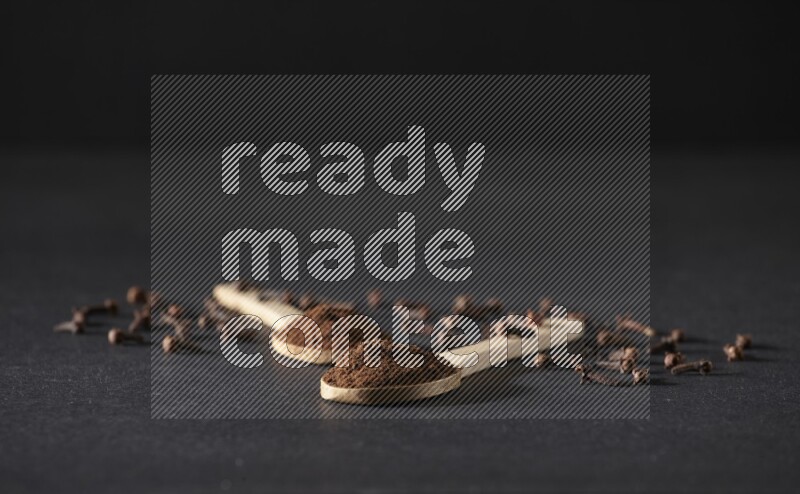 2 wooden spoons full of cloves powder with spreaded whole cloves on a black flooring